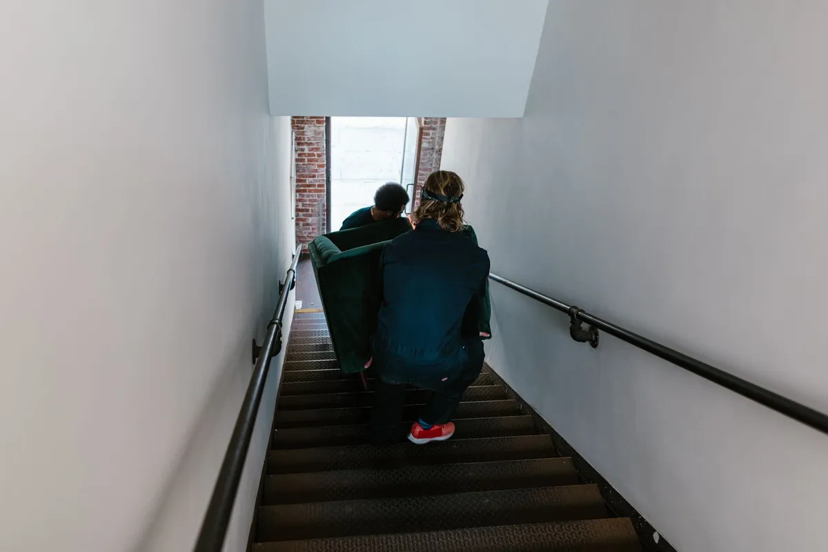Zwei Umzugshelfer tragen ein dunkelgrünes Sofa eine enge Treppe hinunter, von oben fotografiert, Rückenansicht