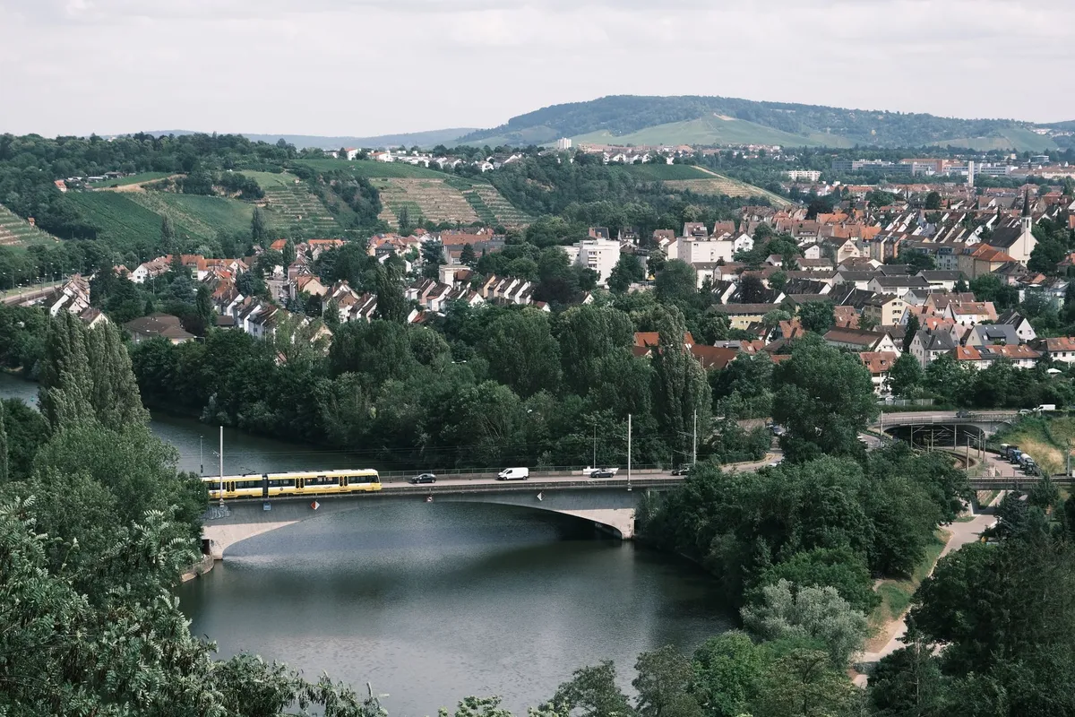 Blick über den Neckar auf eine Brücke mit gelber Stadtbahn, dahinter Wohnhäuser am Hang und Weinberge am Stadtrand