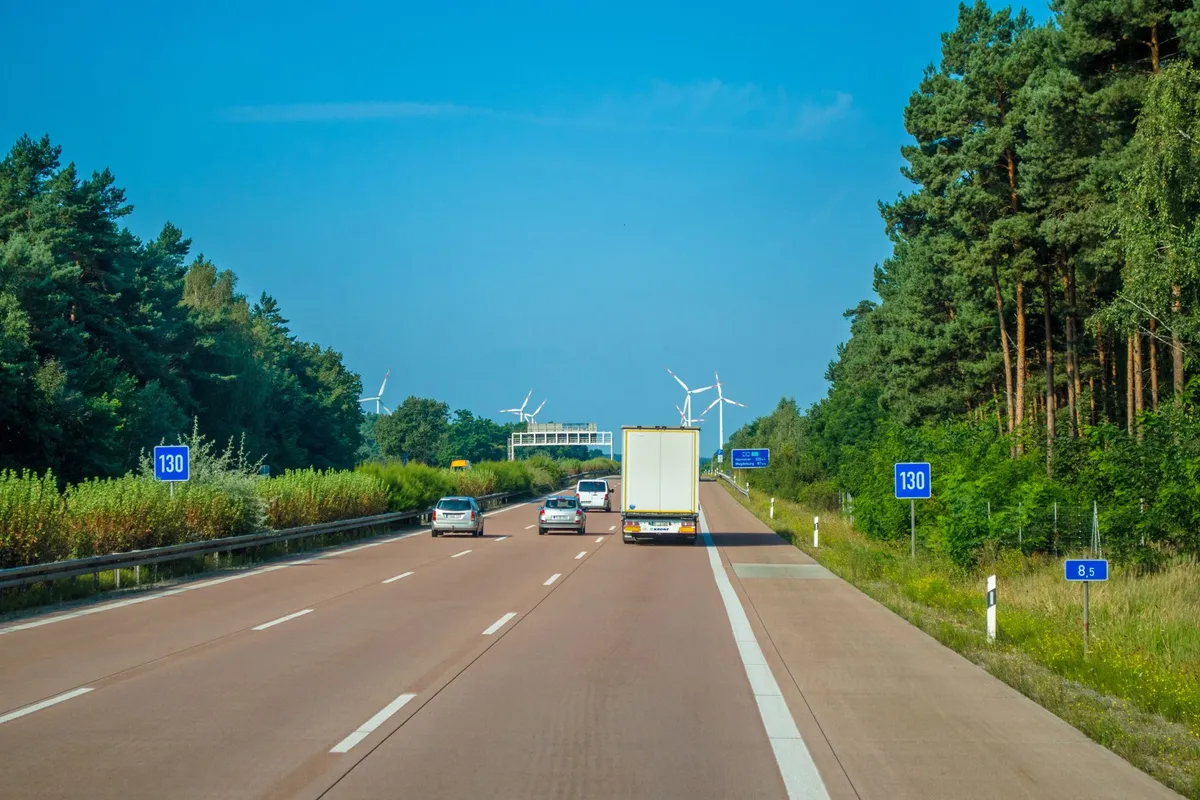 Weißer Lastwagen ohne Firmennamen fährt auf einer deutschen Autobahn, am Rand Kiefern, Windräder und blauer Himmel