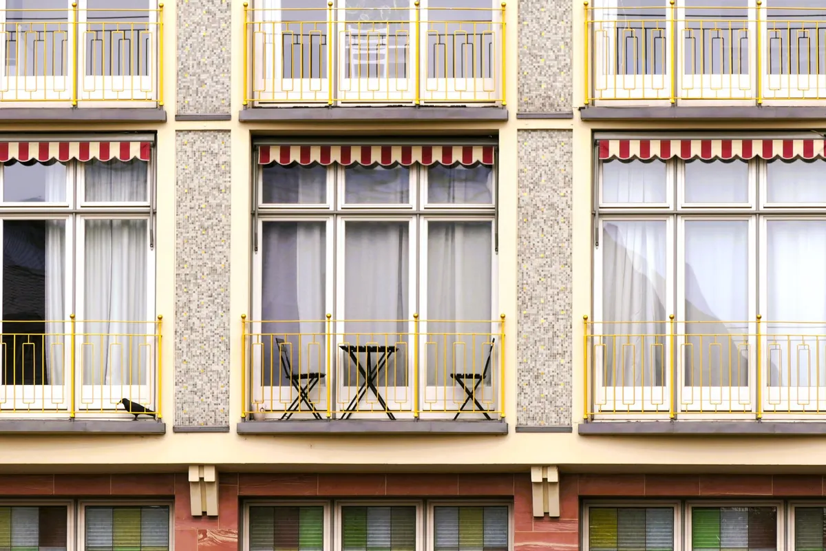 Mehrstöckige Wohnhausfassade mit gelben Balkongeländern, rot-weiß gestreiften Markisen und kleinem Bistrotisch auf dem mittleren Balkon