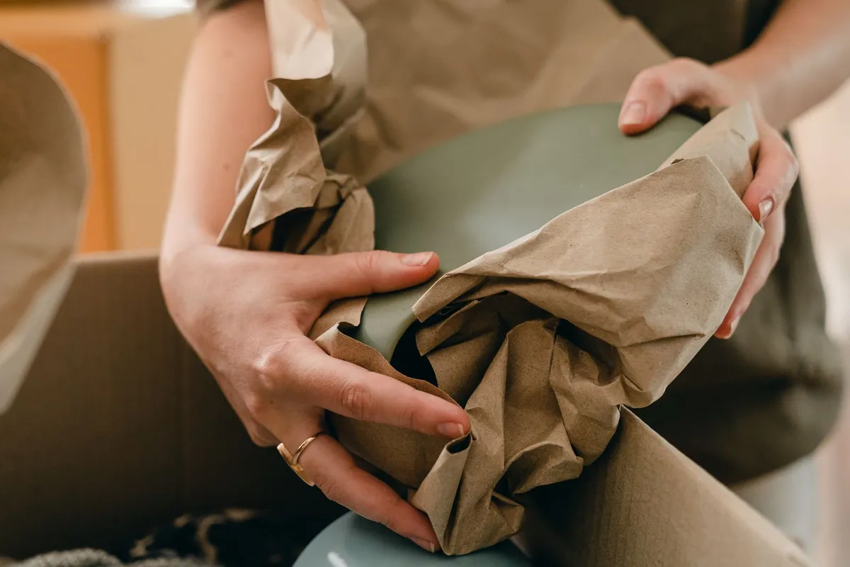 Zwei Hände wickeln einen grünen Keramikteller in braunes Packpapier und legen ihn in einen offenen Umzugskarton