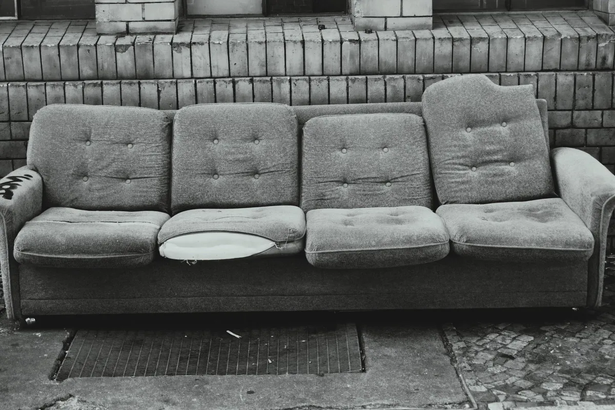 Schwarzweißbild eines ausrangierten Sofas mit aufgeplatztem Sitzkissen auf dem Gehweg vor einer gekachelten Hauswand
